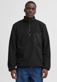 Pullover in pile nero con collo alto, zip a metà e tasca frontale. Superficie testurizzata con vestibilità comoda e maniche lunghe.
