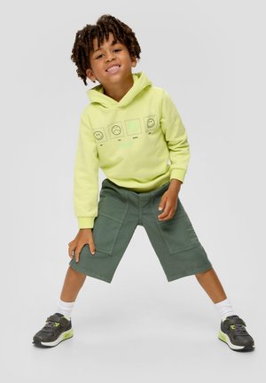 Sweatshirt à capuche jaune vif avec des graphiques de smiley, associé à un short cargo vert et des chaussures de sport noires avec des accents néon.