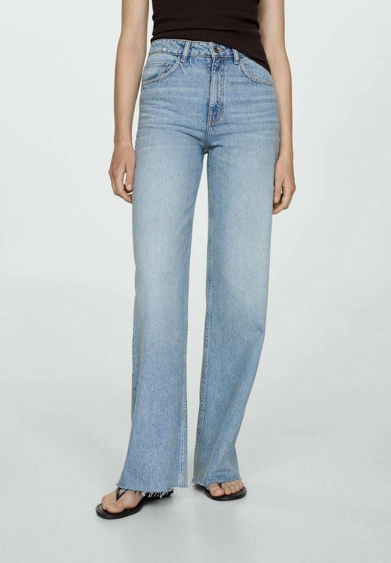 Femme portant un jean large bleu clair avec des ourlets effilochés, un top noir sans manches et des sandales plates noires sur un fond clair uni.