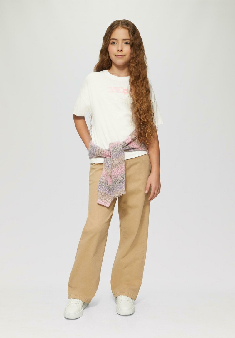Witte t-shirt met roze graphic, beige wijde broek, en een gebreid pastelkleurig sweater om de taille geknoopt. Witte sneakers maken de outfit compleet.