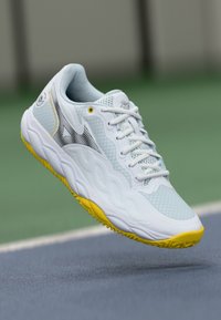 Fehér sportcipő textúrázott hálós felsőrészsel, sárga kiemelésekkel a talpon, és letisztult dizájnnal, fekete logó részlettel.
