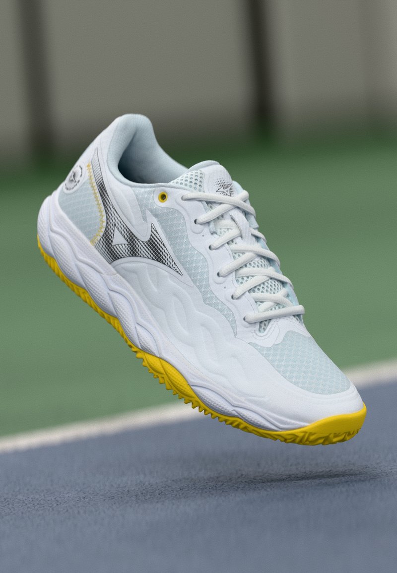 Sapatilha branca desportiva com um upper em malha texturizada, detalhes em amarelo na sola e um design aerodinâmico com um detalhe em logotipo preto.