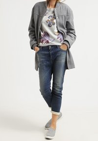 Manteau en maille grise par-dessus un sweat-shirt à motif floral, jean en denim bleu et chaussures grises sans lacets. Tenue décontractée avec des textures superposées.