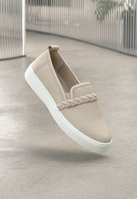 Sapatos slip-on bege com um detalhe trançado na parte da frente, em material de canvas e solas de borracha branca. Design simples com uma textura suave.