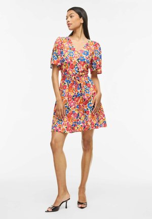 Femme portant une robe fleurie mi-longue avec manches courtes et ceinture à la taille, associée à des mules noires à talons et à bout ouvert, debout devant un fond blanc.