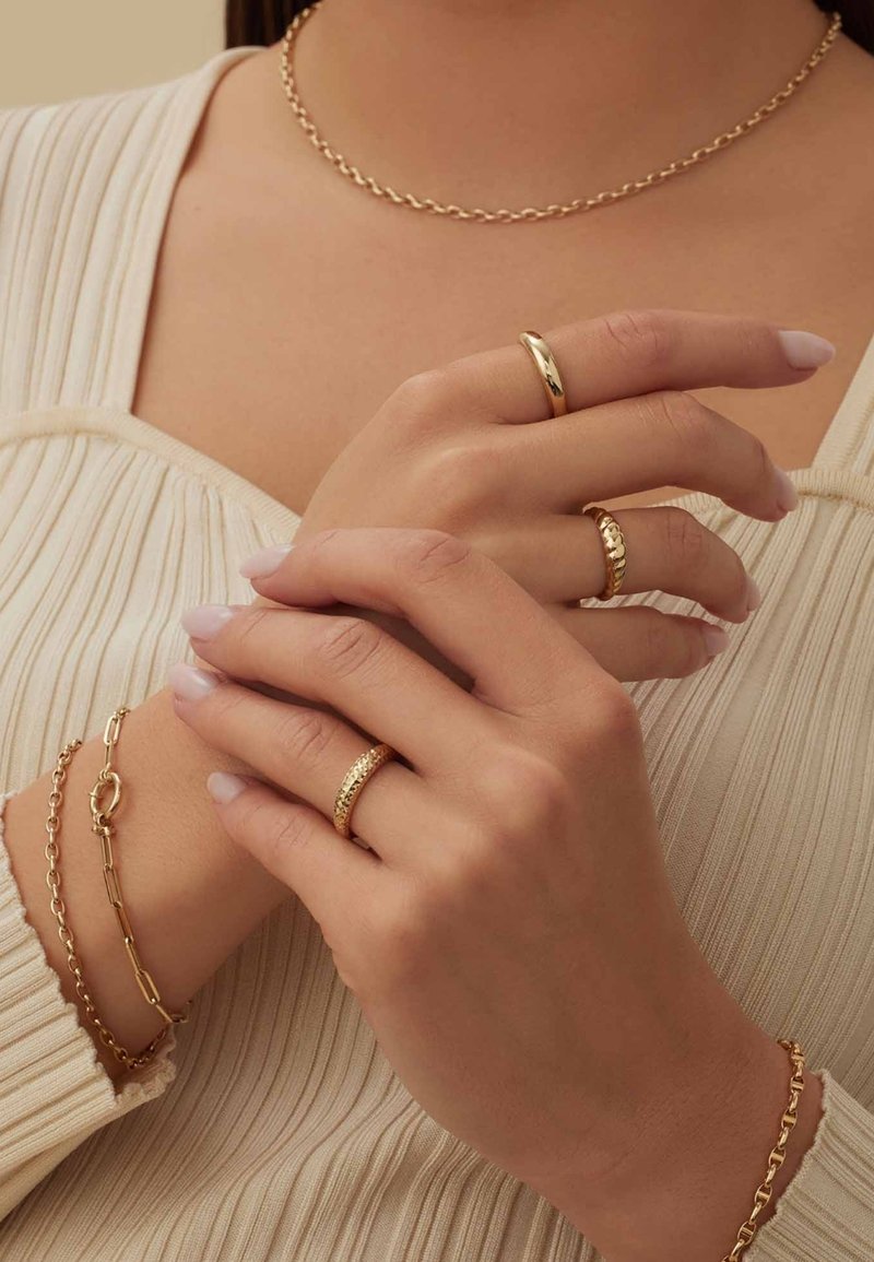 Goldschmuckset, bestehend aus einer zarten Kettenhalskette, drei Ringen mit unterschiedlichen Designs und einem Armband, das alles auf akzentuierten Hauttönen präsentiert wird.