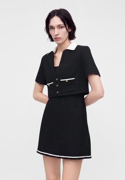 Jeune femme aux cheveux courts et foncés portant une robe noire à manches courtes avec col blanc et bordure, debout devant un fond uni.