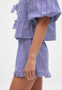 Violet ir baltas gingham stiliaus trumpas viršus su pūstomis rankovėmis, derinamas su atitinkančiomis šortais, turinčiais banguotas kraštines ir vertikalių juostelių raštą.