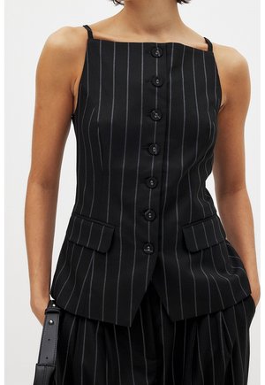 Gilet nero a righe sottili senza maniche con bottoni sul davanti e due tasche con patta, indossato con pantaloni coordinati a righe sottili.