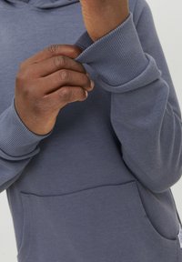 Sudadera gris de tela suave, con puños acanalados y bolsillo. La imagen en primer plano muestra manos ajustando la manga.