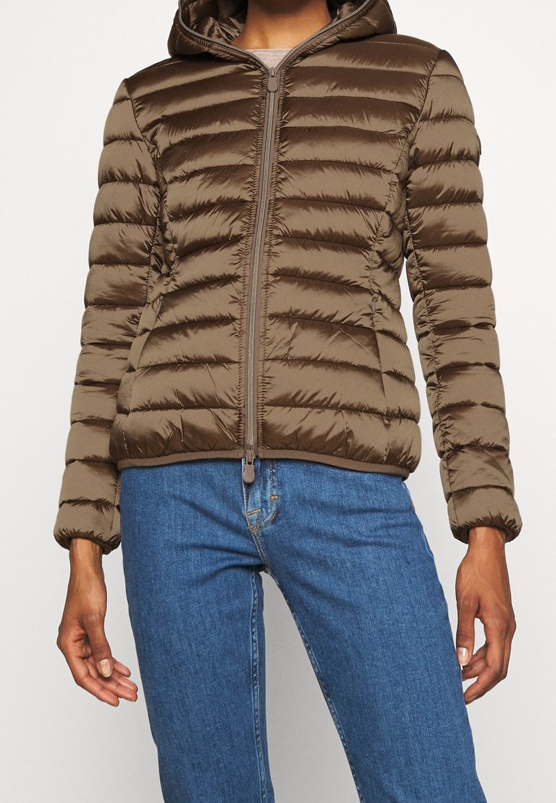 Bruine gewatteerde jas met capuchon, voorzien van horizontale doorgestikte lijnen, ritssluiting en elastische manchetten, gecombineerd met een blauwe spijkerbroek.