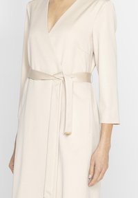 Beżowa sukienka kopertowa z długimi rękawami, z wiązaniem w talii, delikatnym połyskiem i głębokim dekoltem w kształcie litery V. Posiada boczną kieszeń.