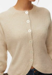 Cardigan beige en maille avec six boutons ronds crème, manches longues et encolure ronde côtelée, porté avec un jean bleu foncé.