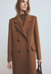 Bruine dubbele rij knopen wollen jas met zwarte knopen, grote revers en zijzakken, gedragen over een donkere trui, met een getailleerde pasvorm.