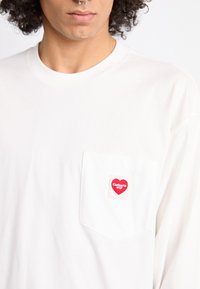 White long-sleeve t-shirt made of cotton, featuring a chest pocket with a red heart logo and "Carhartt WIP" text in white. Soft texture.
