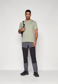 Gaiši zaļa īsroku krekls, pelēkas un melnas bikses, izturīgs dizains, valkāts ar melniem apaviem, rokās tur melnas lences mugursomu.