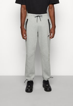 Homme portant un pantalon de survêtement gris clair avec des poches zippées noires, des cordons noirs et des baskets noires, debout contre un mur blanc.