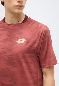 Νεαρό άτομο φορώτας ένα αθλητικό πουκάμισο σε χρώμα μαρόν με σχέδια και με μπεζ λογότυπο στο στήθος, διακομμένο στον λαιμό και το χέρι.