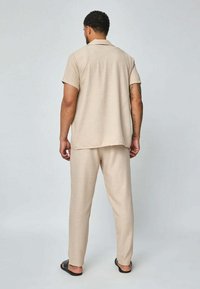 Ensemble de chemise à manches courtes beige et de pantalon assorti, présentant une coupe décontractée et un tissu texturé. Porté avec des sandales noires.