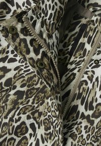 Jaktjacka i leopardmönster i nyanser av brunt och vitt. Har dragkedja och tryckknappar, med en slät yta och mönstrad design.