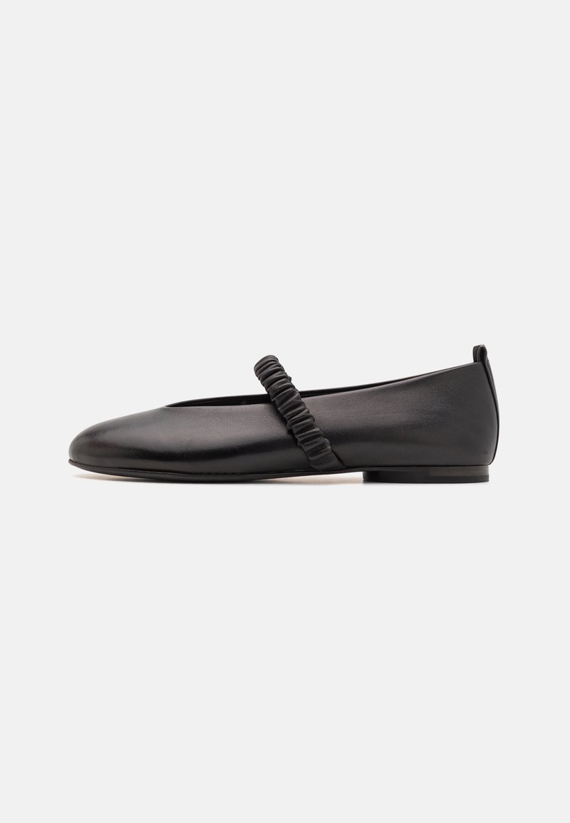 Chaussure plate en cuir noir avec une sangle élastique froncée sur le cou-de-pied et un petit talon sur fond blanc.