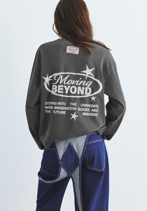 Femme aux longs cheveux portant un sweat-shirt gris foncé avec le texte "Moving Beyond" et des étoiles, associé à un jean bleu avec des patches gris clair.
