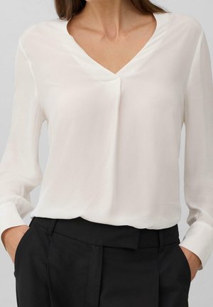 Mujer con blusa blanca de manga larga y cuello en V, metida dentro de pantalones negros a medida, con las manos en los bolsillos, frente a un fondo liso.