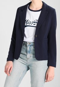 Námořnický modrý blazer s klopovými límci, zapínáním na jeden knoflík a dvěma předními kapsami, nošený přes bílé tričko a světle modré džíny.