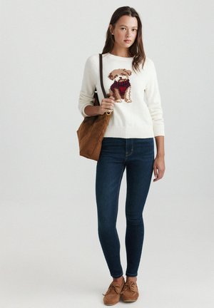 Femme portant un pull blanc avec un motif chien, jean skinny foncé, chaussures couleur sable et sac à bandoulière marron, debout devant un fond uni.