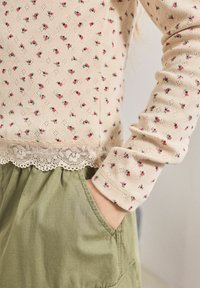 Haut tricot beige clair avec de petits motifs floraux rouges, doté d'un ourlet en dentelle festonné. Associé à un pantalon en tissu vert olive avec poches.