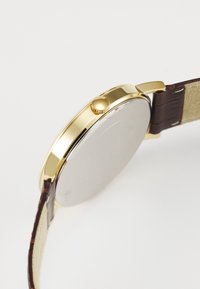Zlatni sat s smešnim kožnim remenom, okruglom kućištem, teksturiranom krunom i glatkom metalnom stražnjom stranom. Elegantni dizajn s minimalnim ukrasima.