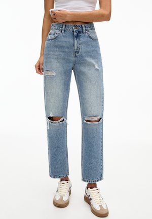 Kvinde iført lyseblå revnede jeans med lige ben og hvide sneakers med beige og brune detaljer, stående mod en hvid baggrund.