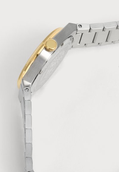 Vue latérale d'une montre-bracelet en métal argenté avec une lunette et une couronne en or, montrant le dos de la montre et le bracelet métallique relié.