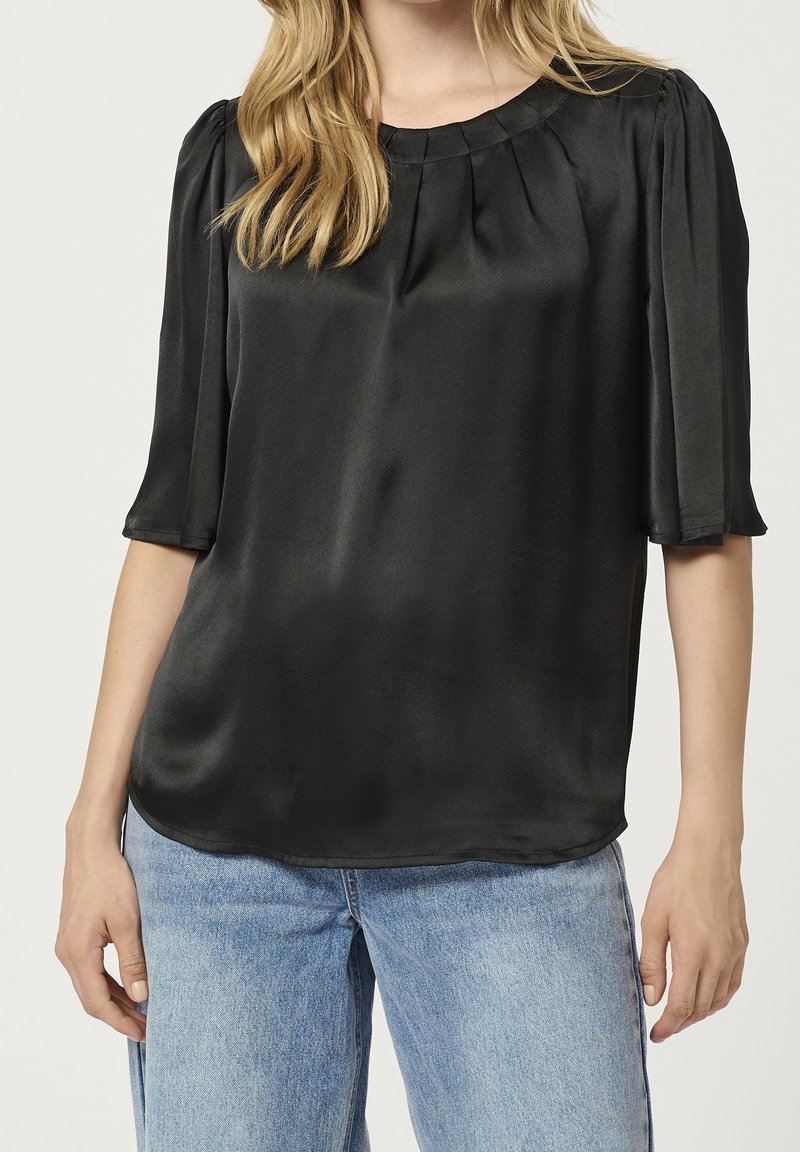 Femme portant un chemisier en satin noir à manches courtes et des plis au niveau du décolleté, associé à un jean bleu clair.