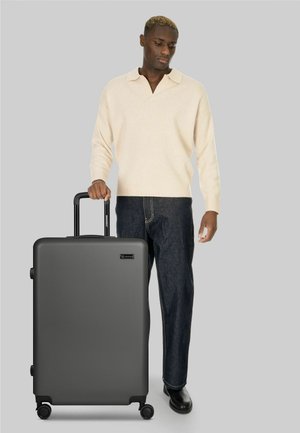 Uomo con maglione beige e jeans scuri in piedi accanto a una grande valigia nera con manico esteso su sfondo grigio.