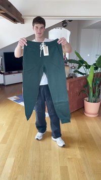 Hombre sosteniendo pantalones de chándal verde oscuro con una etiqueta de cordón blanco en una sala de estar luminosa con suelo de madera y plantas.