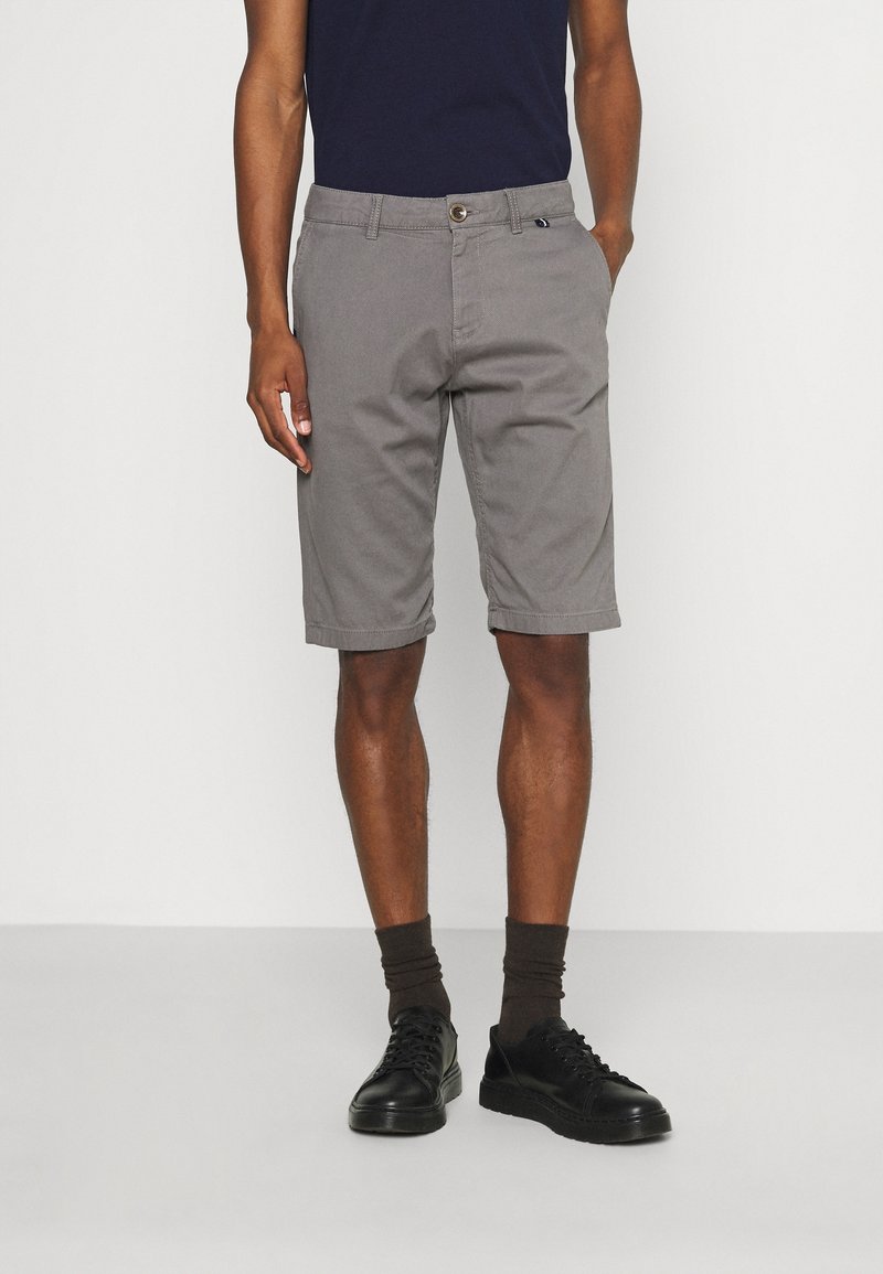 Homme portant un short gris au genou, des chaussettes noires montantes et des chaussures noires à lacets, debout devant un fond blanc uni.
