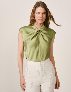 Mujer con blusa sin mangas de satén verde oliva con escote retorcido, combinada con pantalones blancos de talle alto bordados con motivos de hojas.