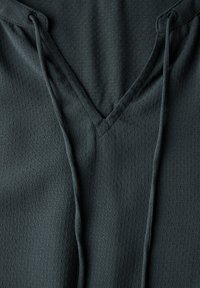 Donkergroene hoodie met een v-hals ontwerp, gemaakt van textuurstof met kleine, verhoogde stippen. Inclusief verstelbare koorden bij de hals.