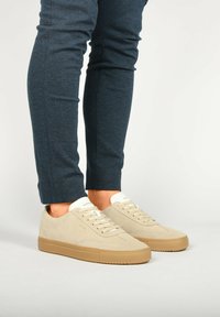 Beige mokkasuohkikengät valkoisilla nauhoilla ja vaalealla kielellä, yhdistetty tummansiniisiin kapeneviin housuihin. Sileä pinta ja minimalinen muotoilu.