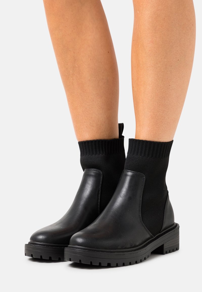 Botines negros hasta el tobillo con acabado en cuero liso, paneles laterales elásticos y puños de tejido texturizado. Suelas gruesas de goma que ofrecen tracción y estabilidad.