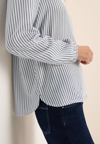 Chemise rayée en bleu clair et blanc, dotée de manches longues et d'une coupe décontractée. Le tissu est léger, avec une texture subtile et un ourlet asymétrique.