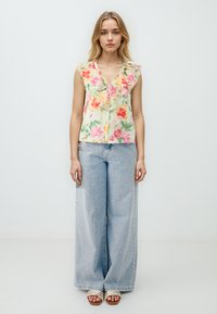 Ärmelloses Blumenshirt aus pastellgelbem Stoff mit rosa und grünen Mustern, das einen gerafften Ausschnitt hat, kombiniert mit hellblauen weiten Jeans.