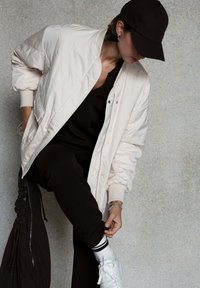 Personne portant une casquette noire, une tenue noire, une veste blanche matelassée, ajustant un pantalon noir par-dessus des baskets blanches devant un mur gris.