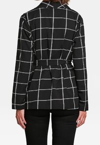 Svart rutigt jacka med vit rutnätmönster, långa ärmar, bälte i midjan och klassisk krage. Slät tyg och strukturerad design.