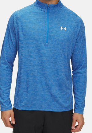 Mann trägt einen blauen, langärmligen Sportpullover mit Viertelreißverschluss und weißem Logo, kombiniert mit schwarzen Hosen.