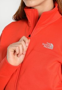 Chaqueta de forro polar roja con cuello alto, cremallera frontal y el logo de The North Face en plata. Textura suave y diseño ajustado.