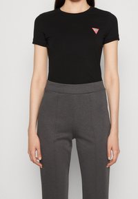 Černé tričko s krátkým rukávem vyrobené z měkké látky, s červeným trojúhelníkovým logem na levém prsu. V kombinaci se šedými kalhotami s vysokým pasem.