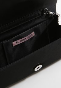 Cartera de mano negra con interior de satén, que presenta un compartimento principal, un cierre a presión plateado y una etiqueta de tela rosa que dice "ANNA FIELD."