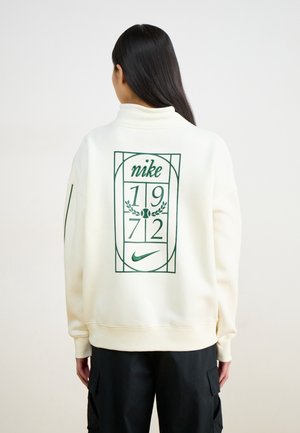 Personne aux longs cheveux noirs portant un sweat-shirt blanc Nike avec un motif vert 1972 et un pantalon noir, vue de dos sur fond uni.